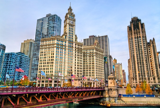 A scenic view of downtown Chicago showcasing skyscrapers, including the iconic Wrigley Building with its clock tower, alongside a bridge over the Chicago River, adorned with flags and autumn foliage.