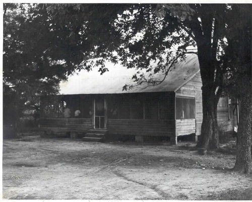 A black and white photograph of a wooden house with a porch, surrounded by trees and a dirt pathway.