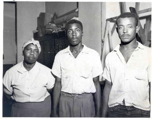 A black and white photo of three individuals standing together. On the left is a woman wearing a headscarf and a blouse, while in the center is a young man with a serious expression in a white shirt. To the right is another young man in a slightly unbuttoned shirt, also looking serious. They are in an indoor setting with blurred background elements.