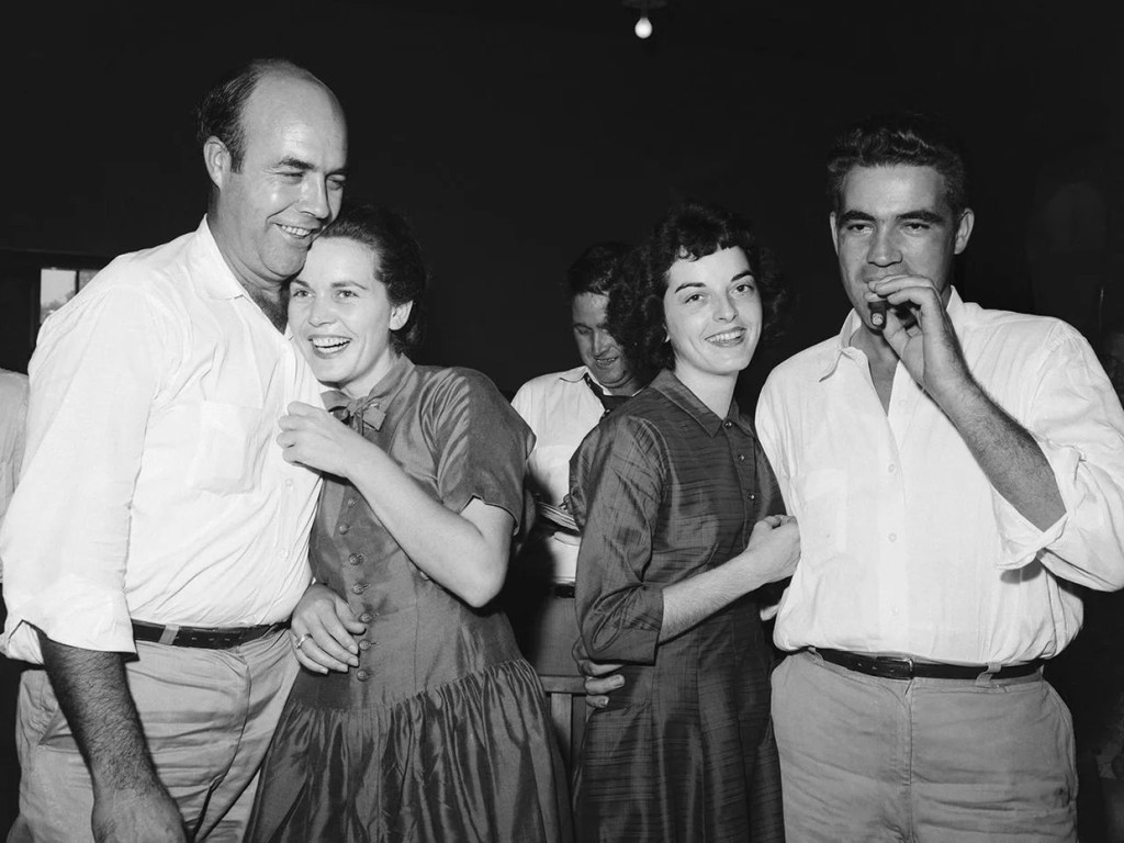 A group of four people joyfully interacting at a social gathering, with two men and two women, one man holding a cigar, and the women smiling and wearing dresses.