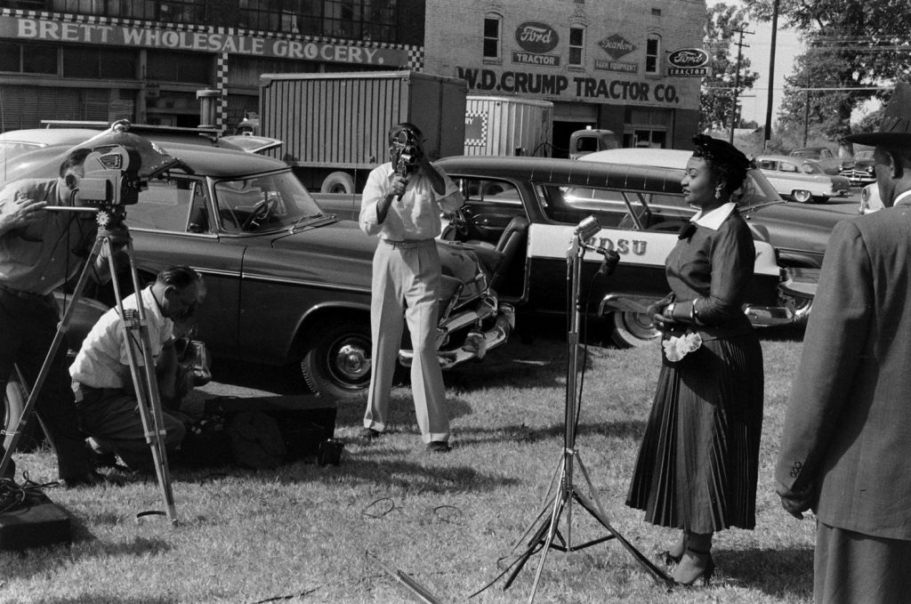 A woman in a formal dress stands at a microphone while several men operate cameras and equipment in a busy outdoor setting with parked vintage cars in the background.