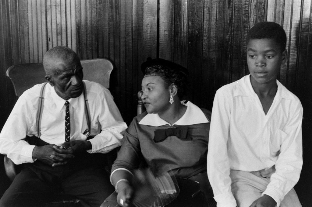 A black and white photograph featuring an elderly man in a suit and suspenders, a woman in an elegant dress with a large bow, and a young boy wearing a white shirt, sitting closely together against a wooden backdrop.