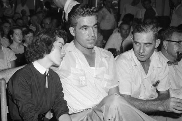 A black and white image of a mixed group of people seated in an audience, featuring a woman leaning against a man in a white shirt, while another man in a light-colored shirt sits beside them, all engaged in the event.