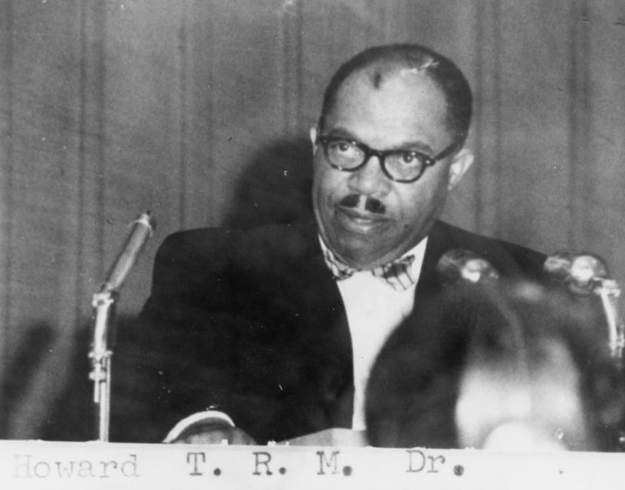 Portrait of a man wearing glasses and a bow tie, seated at a table with microphones, appearing serious.
