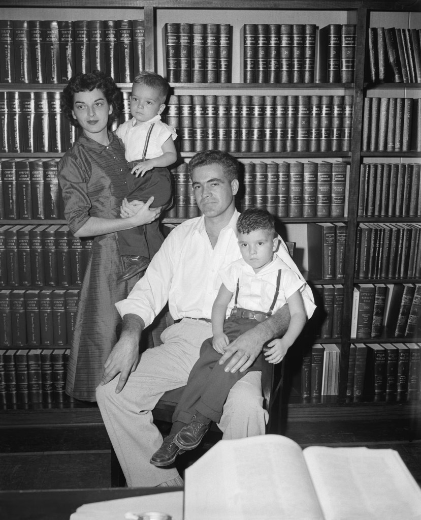 A family portrait featuring a woman holding a young boy, a man seated with another boy on his lap, all in front of a bookshelf filled with books.