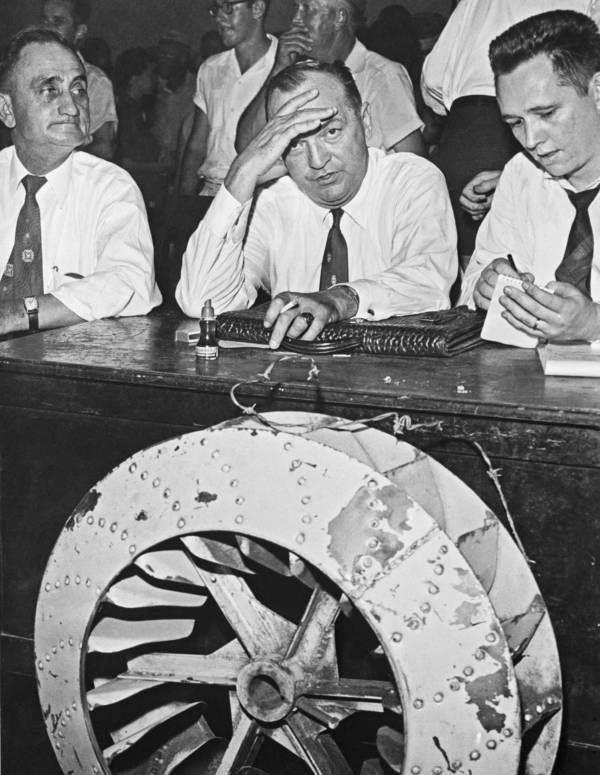 Three men sit at a table, with a large airplane propeller blade visible in the foreground. One man has a concerned expression, resting his forehead on his hand, while the others appear focused on an item on the table.