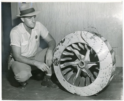 A man in a hat kneels beside a large, circular aircraft part with multiple blades and a metallic surface, inspecting it.