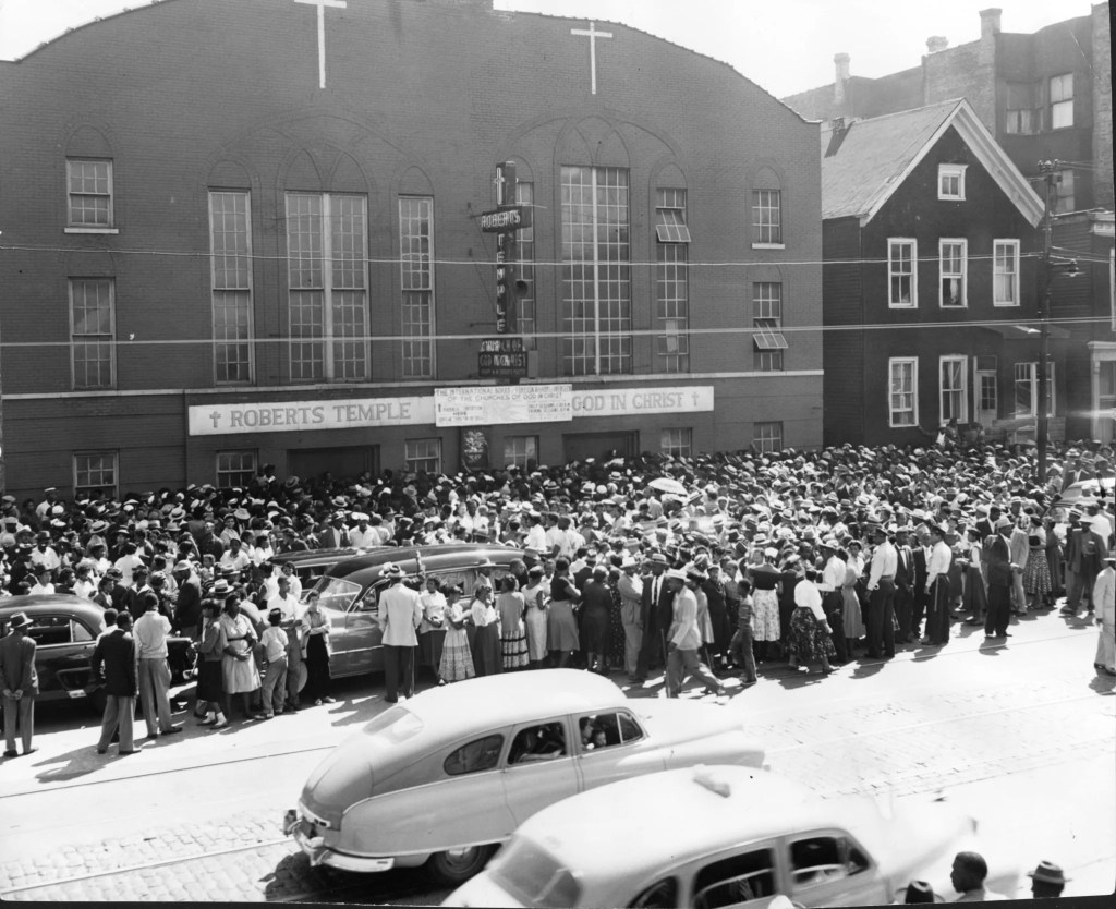 Crowd gathered outside Roberts Temple, with people in hats and formal attire, and vintage cars in the foreground.