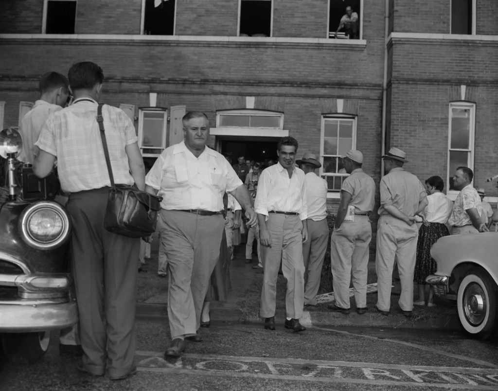 A group of people congregating outside a building, with some individuals walking away, and others standing in a line, while two vintage cars are parked nearby.