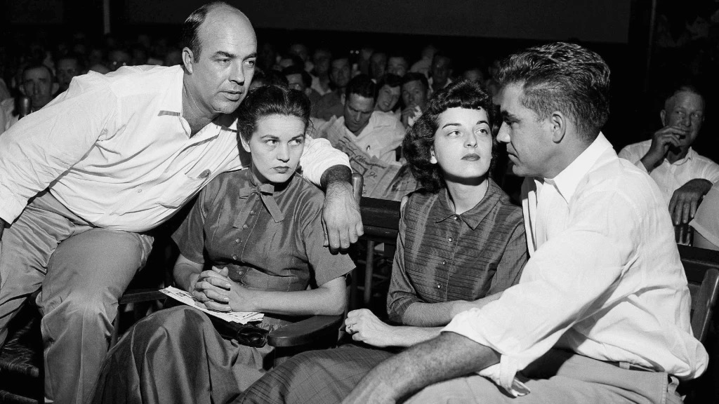 A black and white photograph of four people engaged in conversation in a crowded setting. Two men are leaning towards two women, who are seated. The expressions of the individuals suggest a serious discussion.