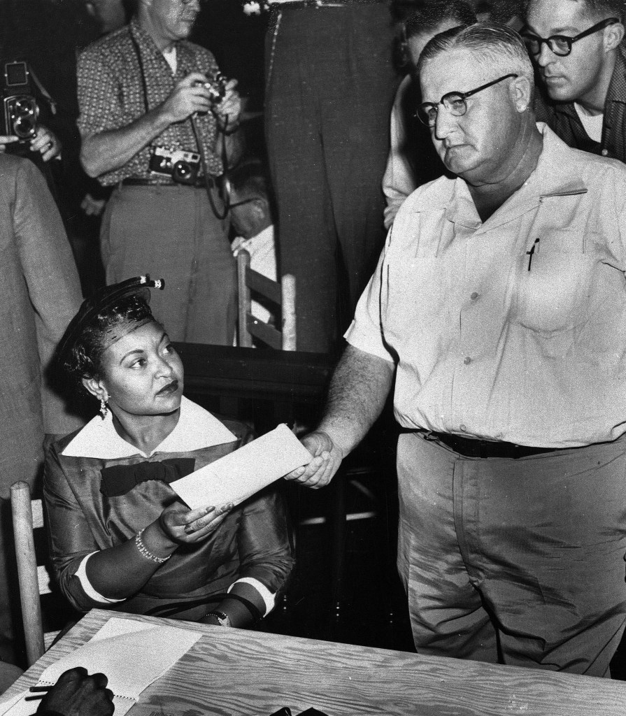 A woman sitting at a table, holding a piece of paper, while a man in a white shirt hands her another document. Several people with cameras are seen in the background.