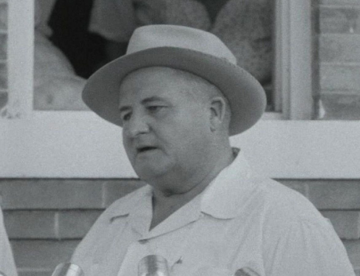 Black and white image of a man wearing a wide-brimmed hat and a collared shirt, speaking at a podium surrounded by microphones, with a window frame in the background.