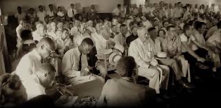 A large crowd of people gathered in a room, with some individuals seated at a table in the foreground taking notes, while others attentively listen in the background.