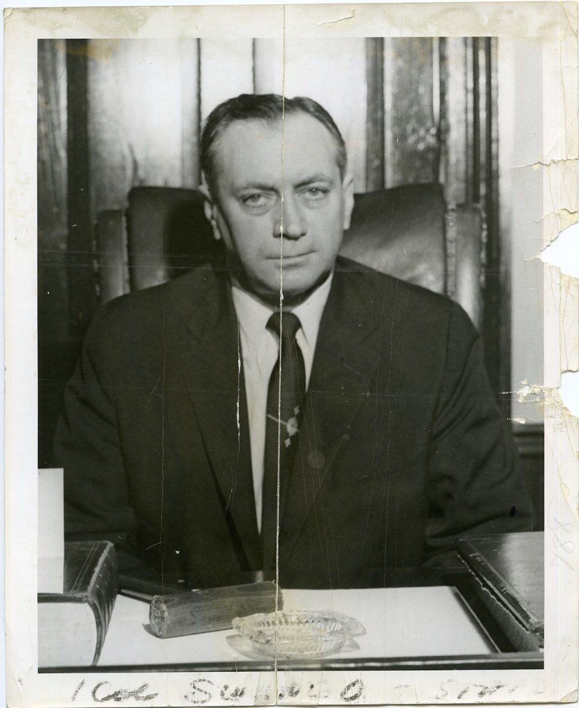 Black and white portrait of a serious man sitting at a desk, wearing a suit and tie, with a wooden backdrop and various items including a book and an ashtray on the desk.
