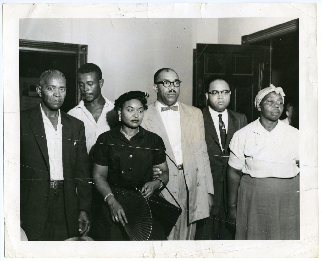 A black and white photograph featuring six individuals standing together, looking serious. The group includes three men and three women, dressed in formal and semi-formal attire. One woman is holding a fan, and they are positioned in an indoor setting with doors in the background.