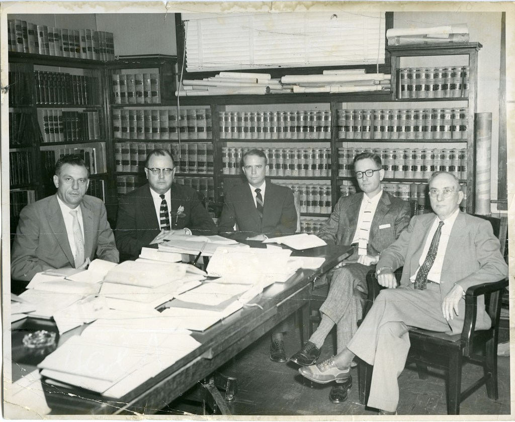 Black and white photograph of five men sitting around a large table covered with papers, with shelves of legal books in the background.